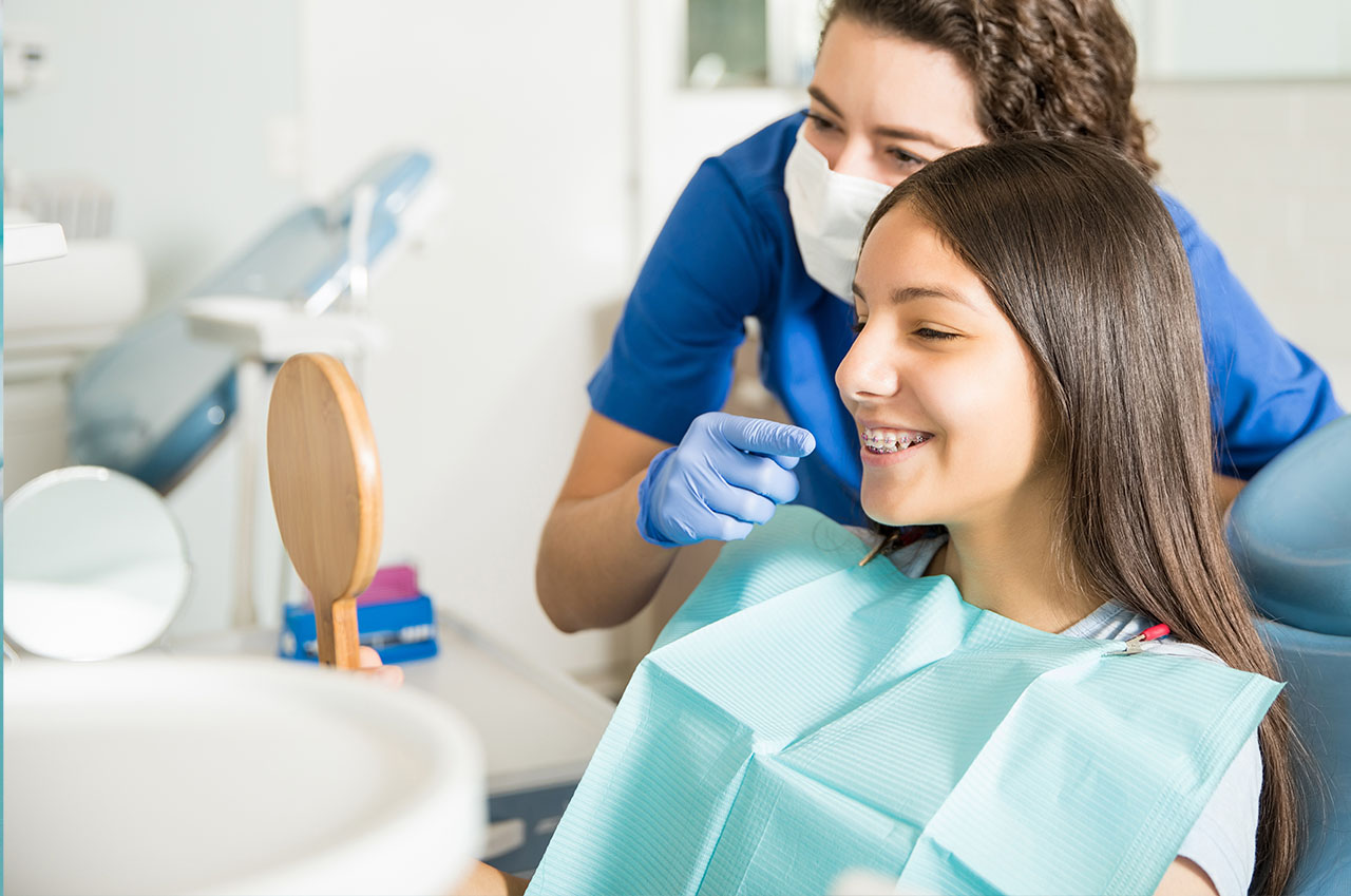 The image shows a dental hygienist assisting a patient during a teeth cleaning appointment, with the dental hygienist wearing protective gloves and using dental tools while the patient sits in the dental chair.