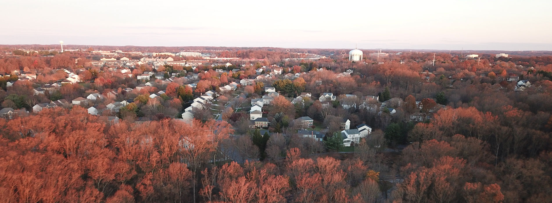 The image shows a panoramic view of a residential area during sunset with red autumn foliage on the trees, capturing the transition from day to night in an urban setting.