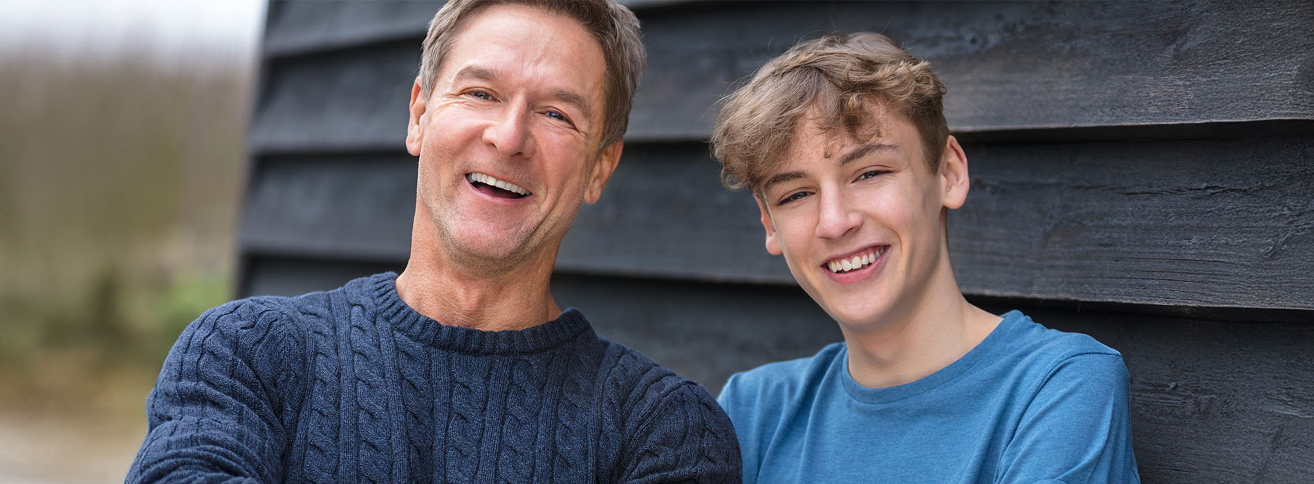 An elderly man and a young boy are smiling at the camera, standing close together with a building behind them.