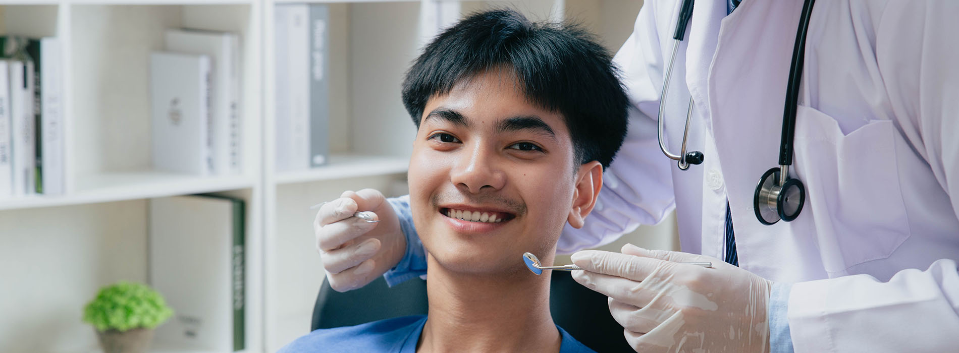 The image shows a young man sitting in a dental chair with his eyes closed, receiving dental treatment from a dentist who is smiling at him.