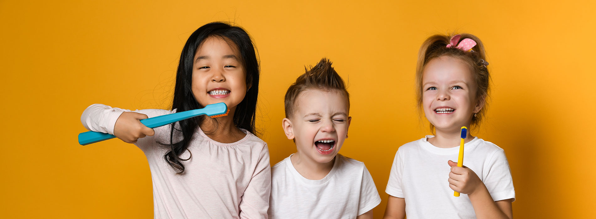 Three children smiling and holding toothbrushes in front of a yellow background.