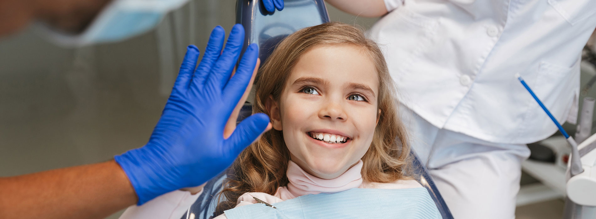 The image shows a young girl with blonde hair sitting in a chair, smiling at the camera, while receiving medical treatment from a healthcare professional wearing a blue gown and gloves, with a hospital setting visible in the background.