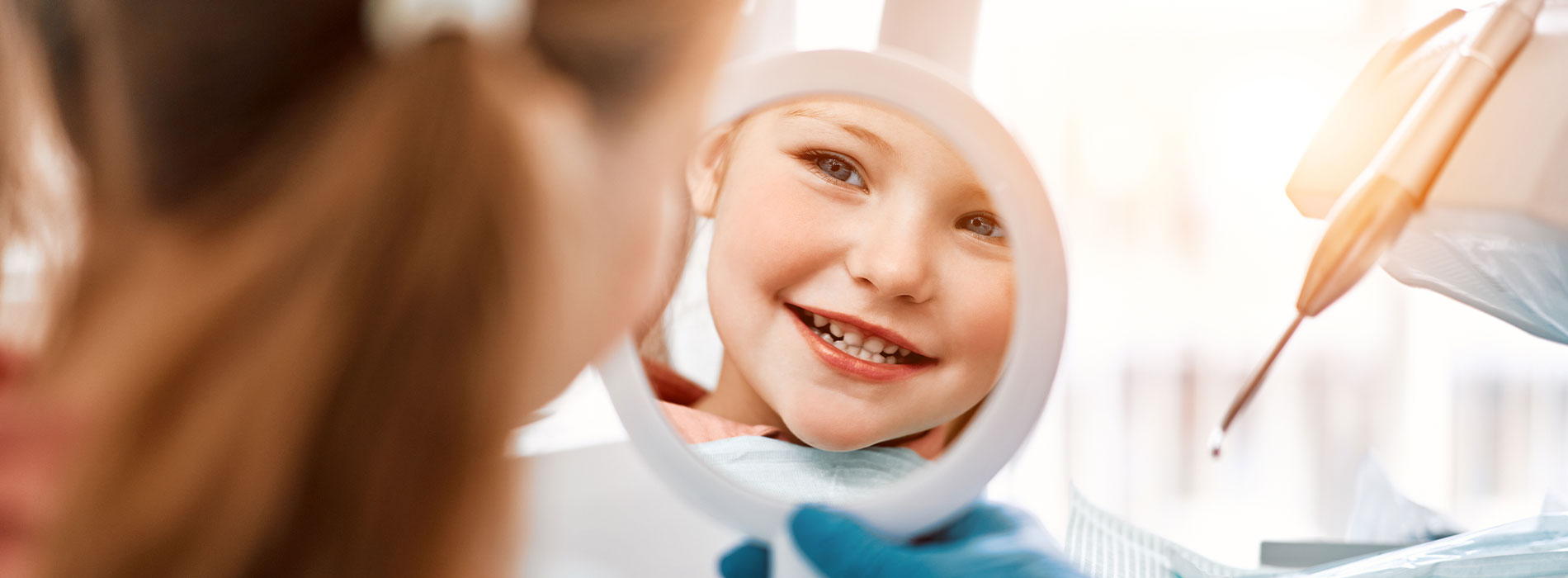 The image shows a young child looking into a mirror with a reflection of their face, with an adult s hands visible in the foreground holding a bottle and a brush, suggesting grooming or beauty care activity.