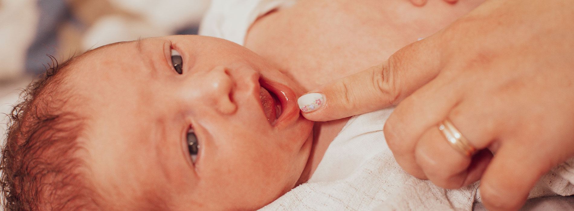 The image shows a newborn baby being held by an adult, with the baby s mouth open and the adult s hand gently placed on the baby s chest.