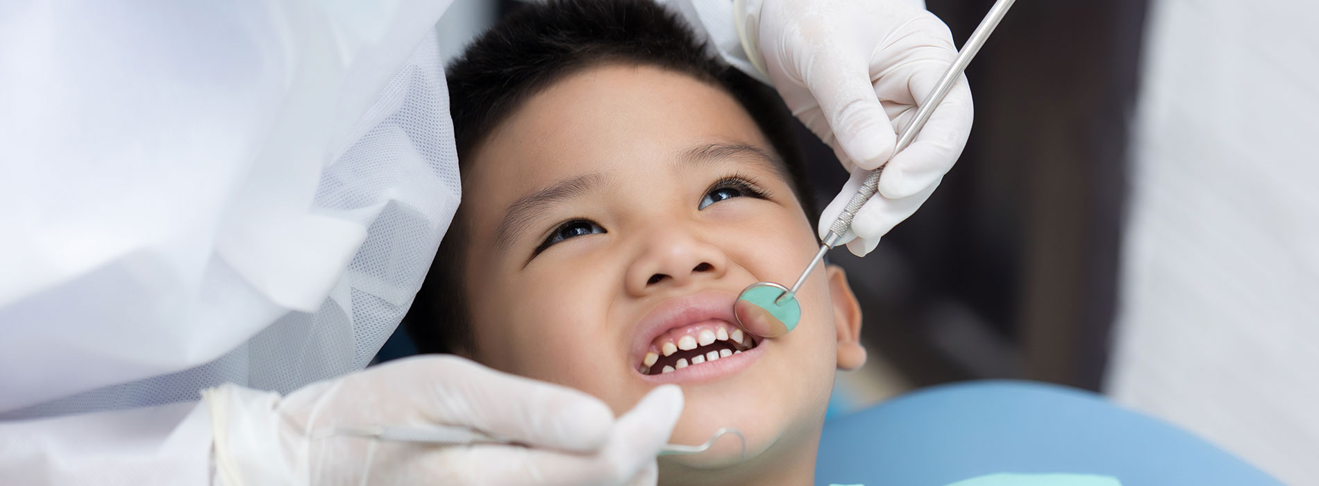 A young boy with dark hair is sitting in a dental chair, smiling broadly while undergoing a dental procedure.