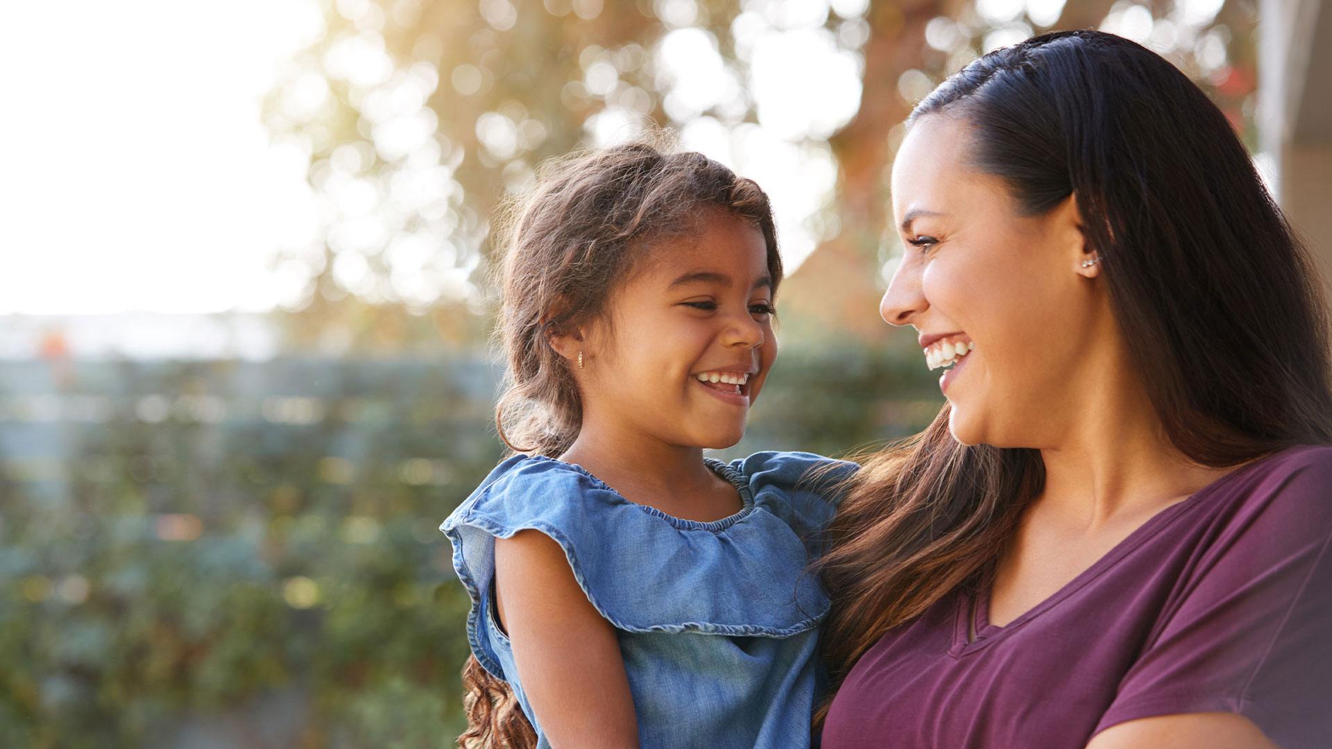 The image shows a woman and a young child smiling at each other outdoors during daylight.