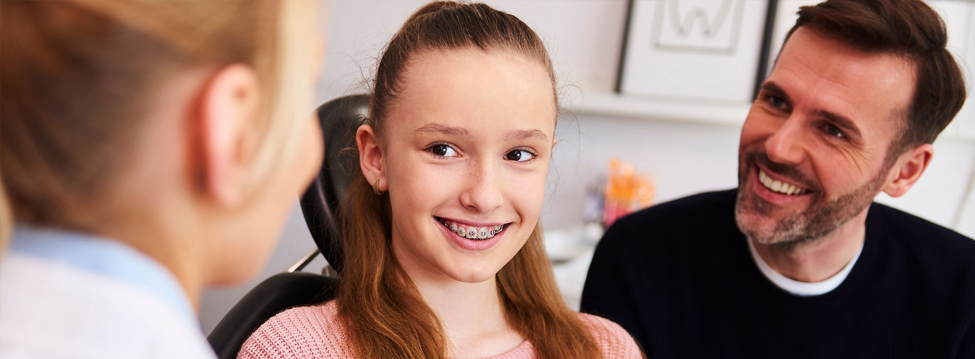 The image shows a young girl smiling at a man who is sitting beside her  another man stands behind them, looking towards the camera with a slight smile. They are indoors, and there s a blurred background that suggests an informal setting.
