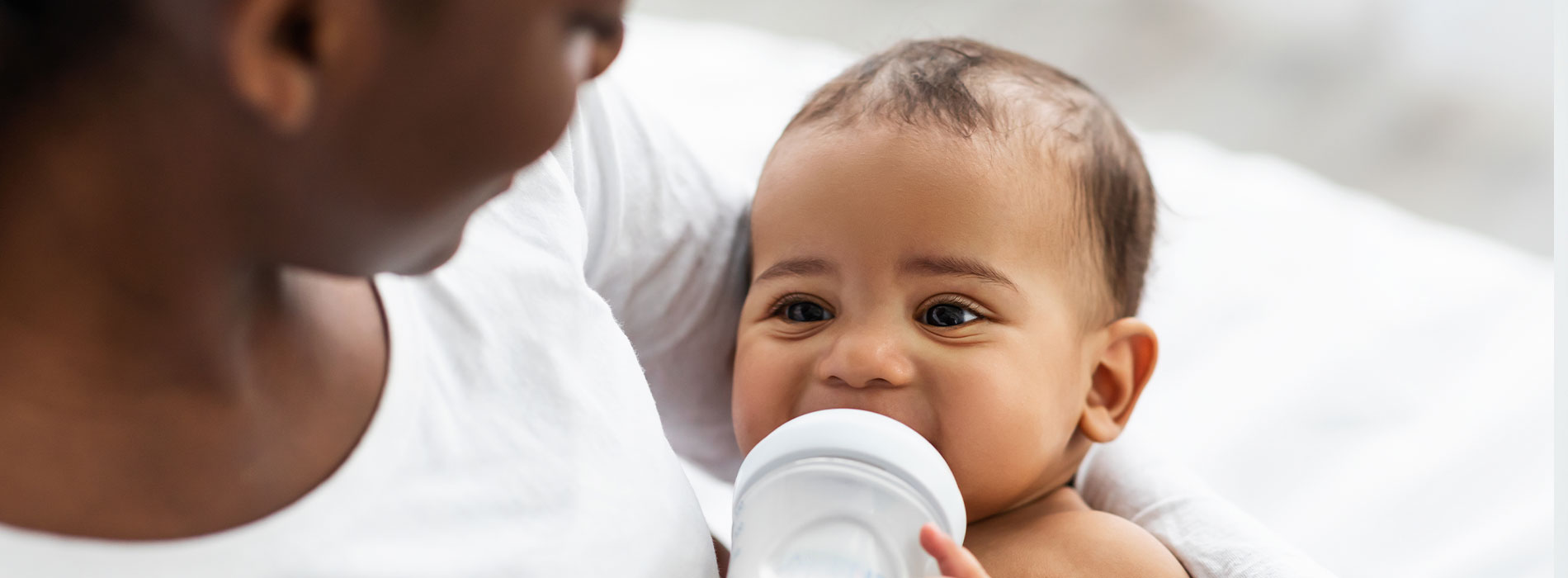 This is a photograph showing an adult holding a baby while looking at the camera, with both individuals appearing to be indoors. The adult s face is partially visible, but the focus is on the child who is smiling and looking towards the camera, with the infant s eyes open wide.