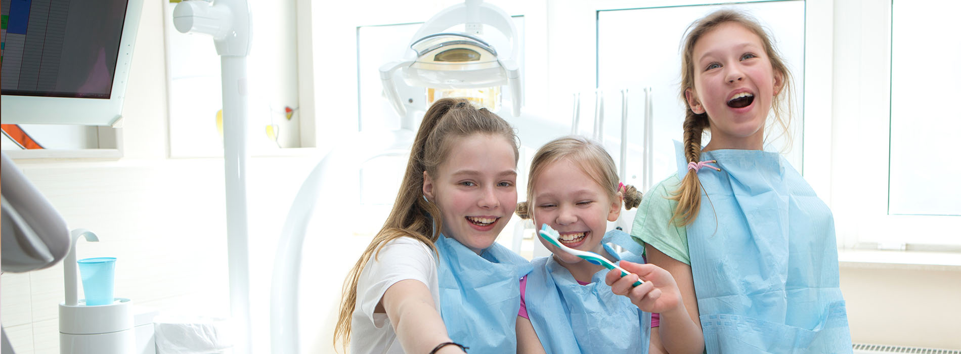 Three young women wearing blue aprons and white face masks, smiling at the camera while posing with dental equipment in a dental office setting.