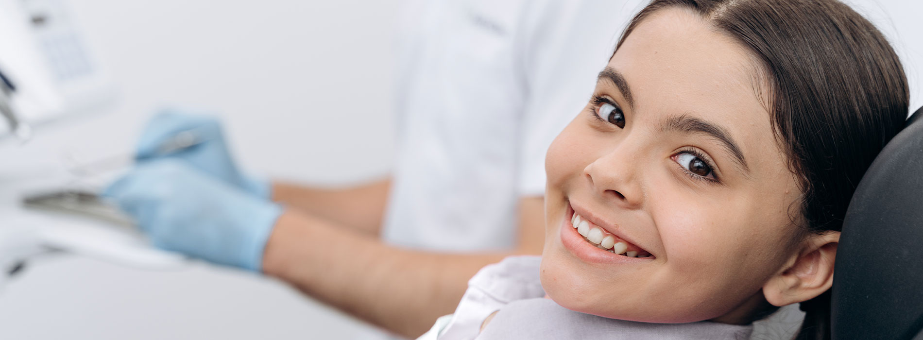 The image shows a young girl with a broad smile, seated in a dental chair, wearing a blue surgical gown, while a dentist works on her teeth, with a dental drill visible in the background.