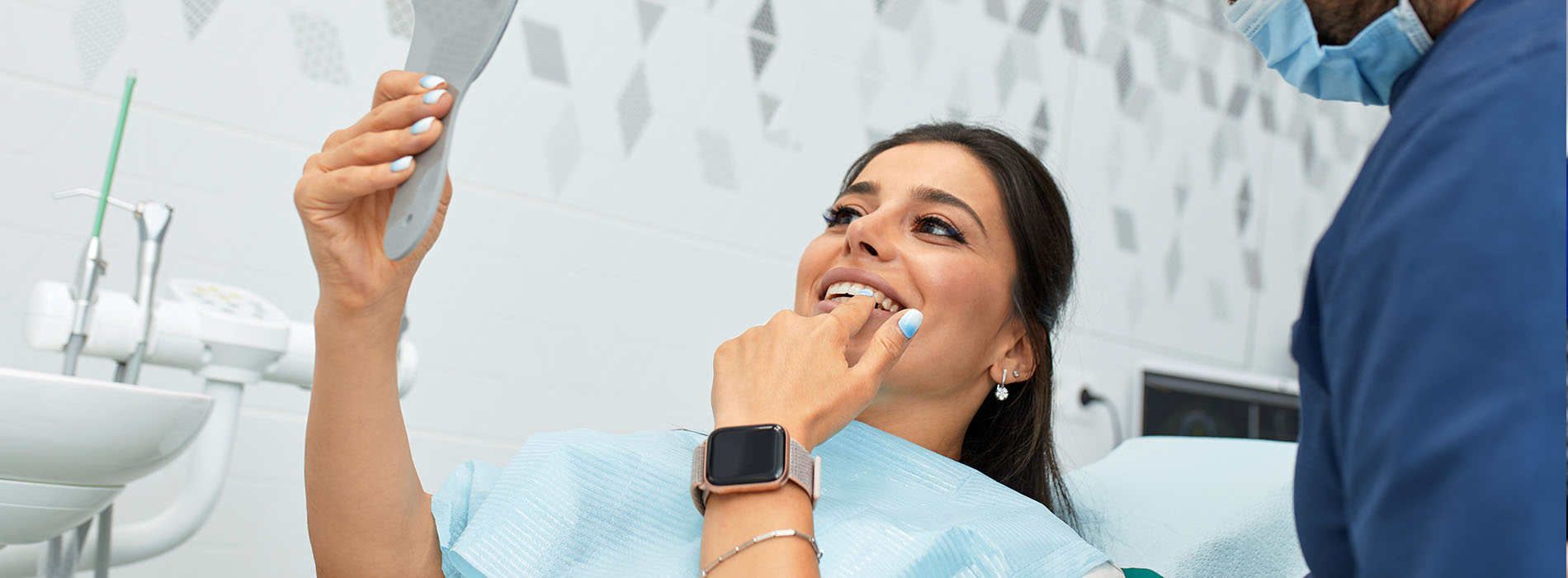 In the image, there is a woman sitting in a dental chair with a dentist standing next to her, holding up a mirror while she brushes her teeth.