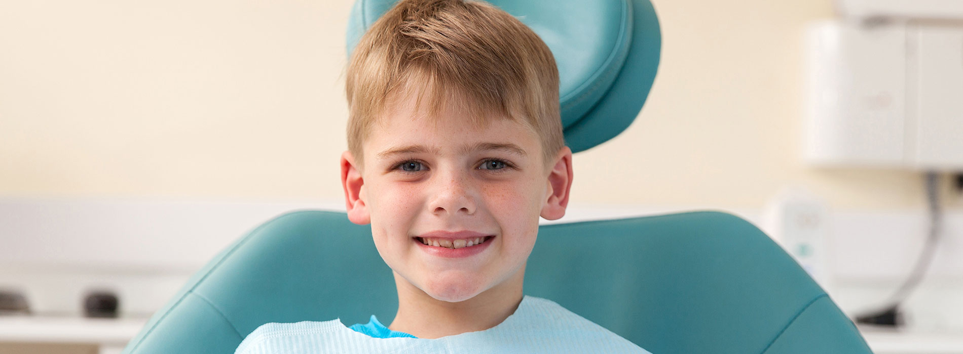 The image shows a young boy sitting in a dental chair, with a blue background and a watermark on the bottom right corner.