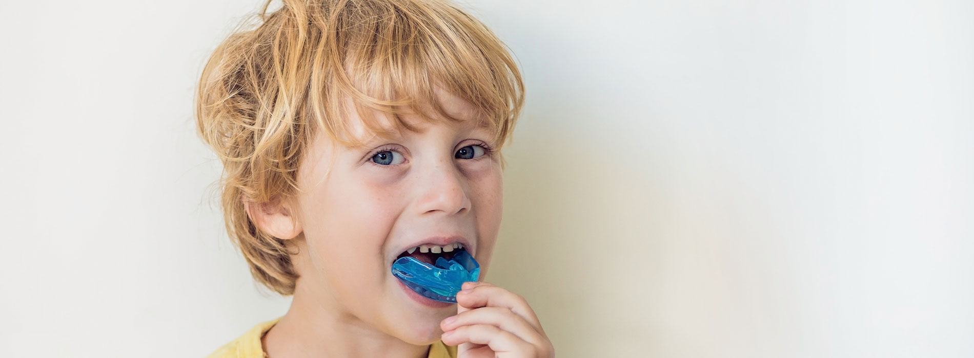 A young child with blonde hair, wearing a yellow shirt, is shown eating a bright blue lollipop.