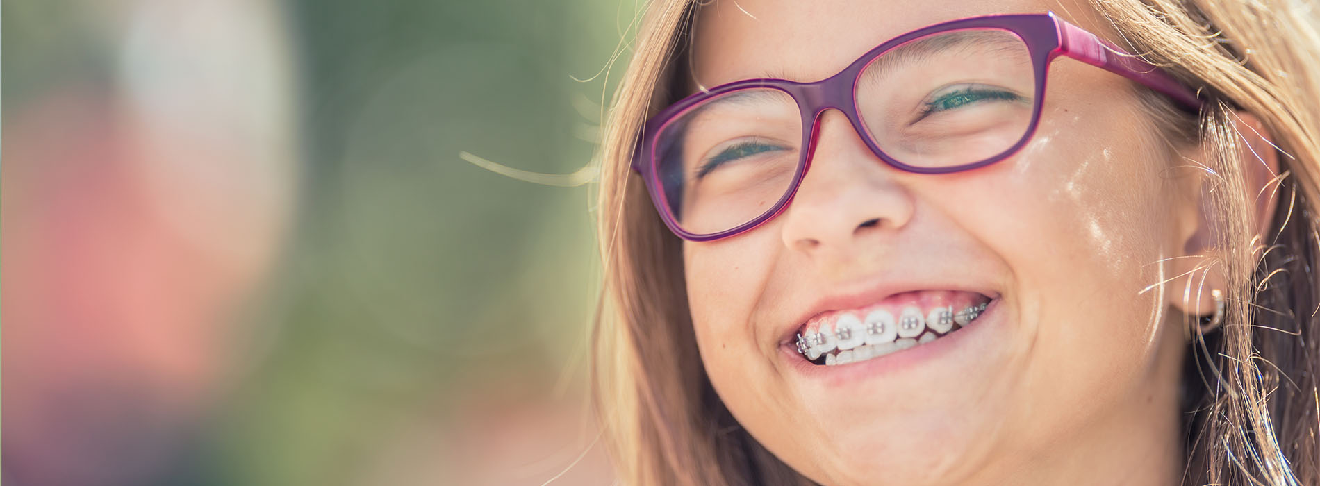 The image shows a young girl with glasses smiling at the camera, with her eyes closed, against a blurred background.