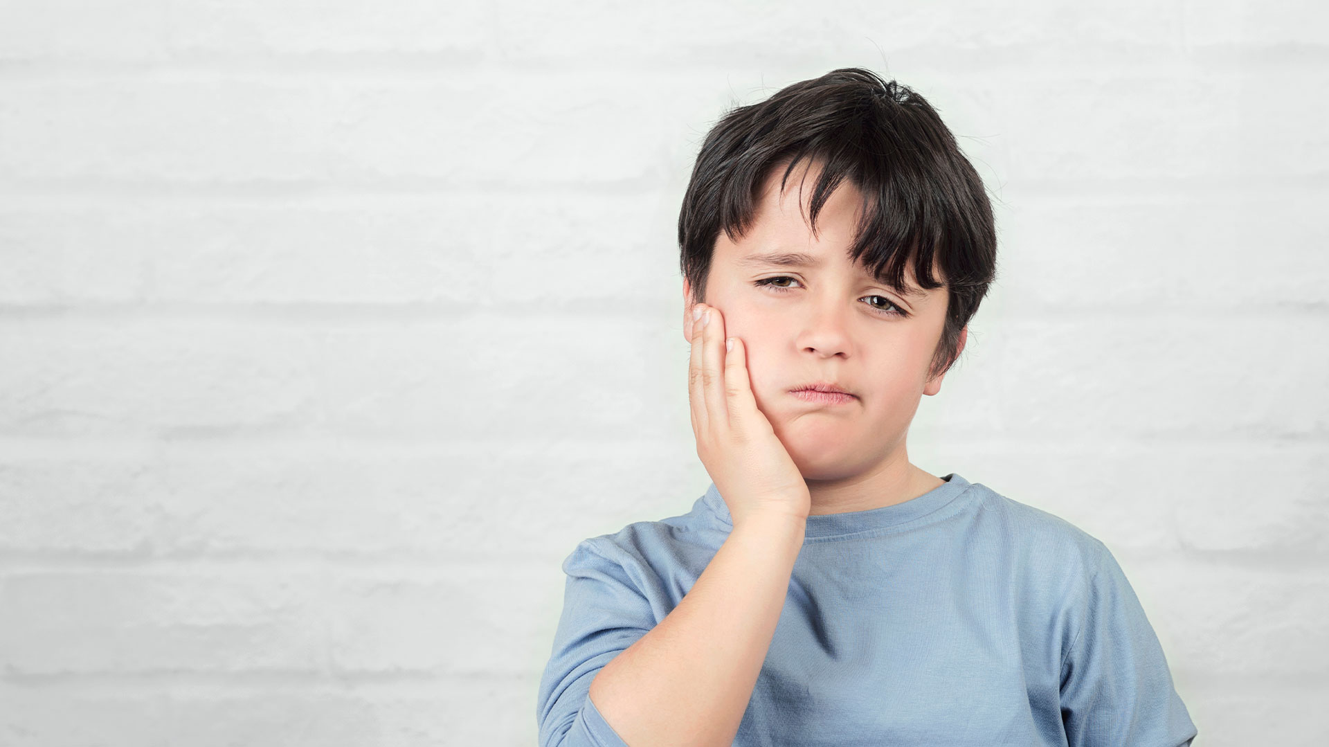 A young boy with his hand on his face, looking directly at the camera, against a plain background.