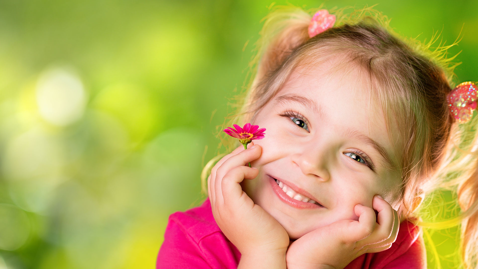 A young girl with rosy cheeks smiles at the camera while posing for a portrait against a blurred background of greenery, with a soft focus on her face.