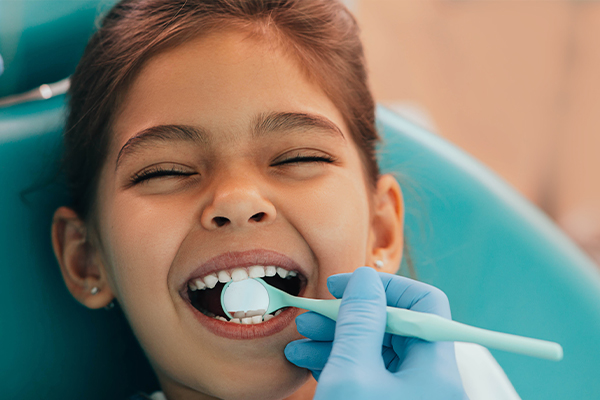 A young girl sitting in a dental chair with her mouth open, receiving dental care, while wearing a blue surgical mask.
