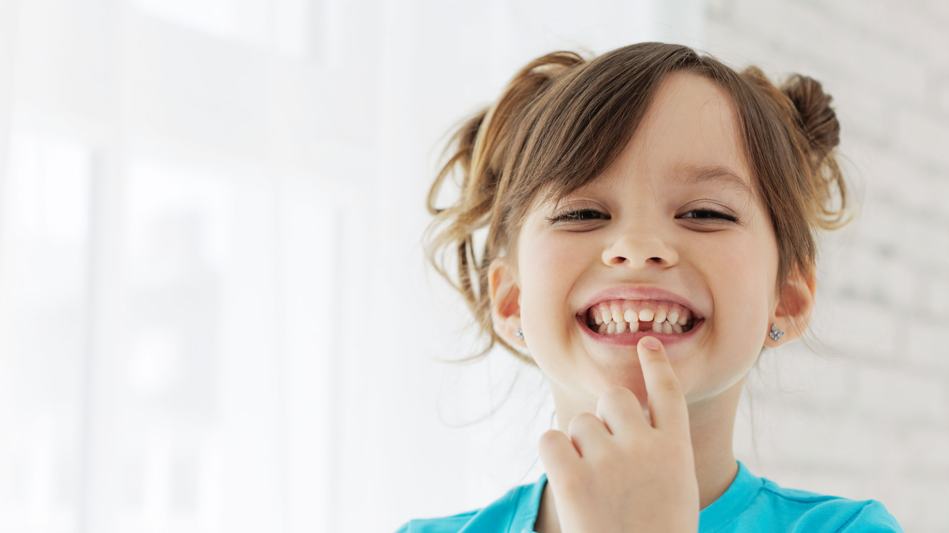 A young girl with her finger in her mouth, smiling at the camera.