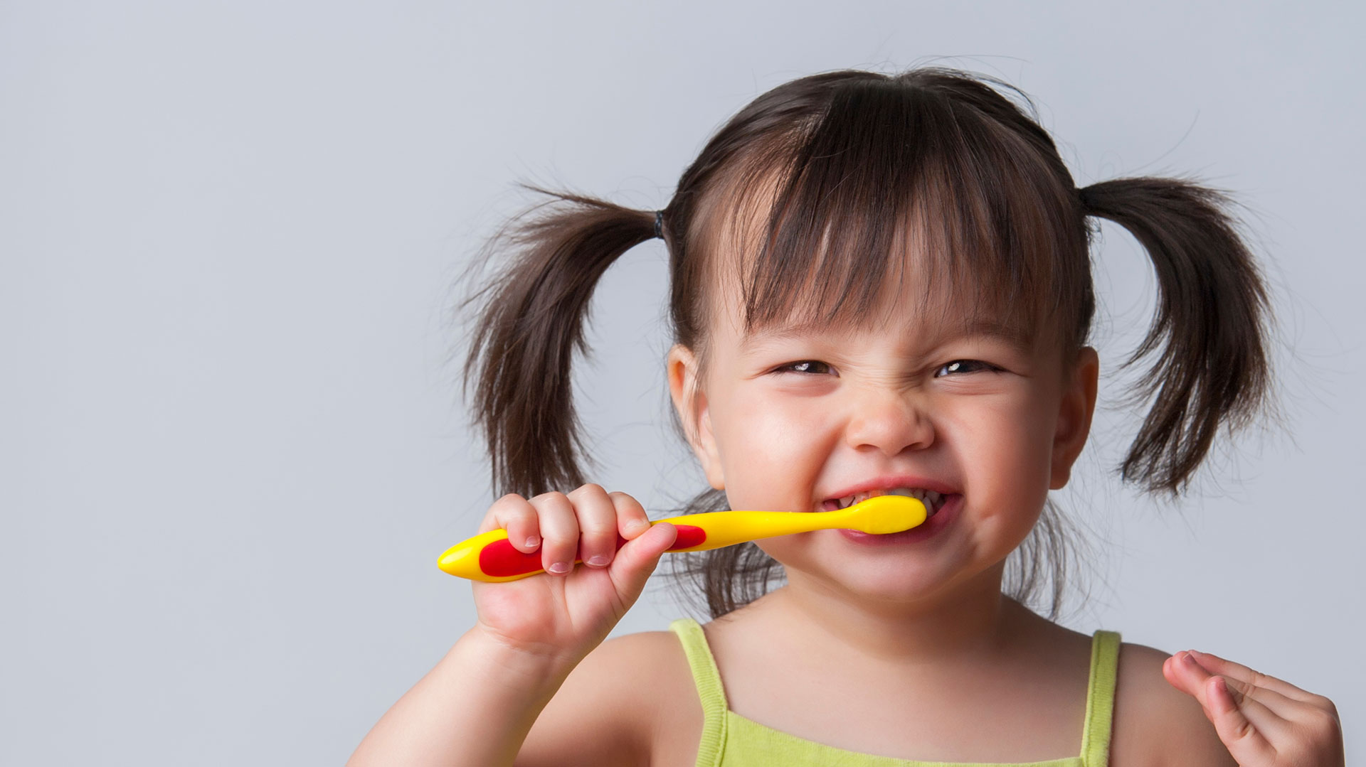 A young child with pigtails is brushing their teeth while making a playful face with a toothbrush in their mouth.