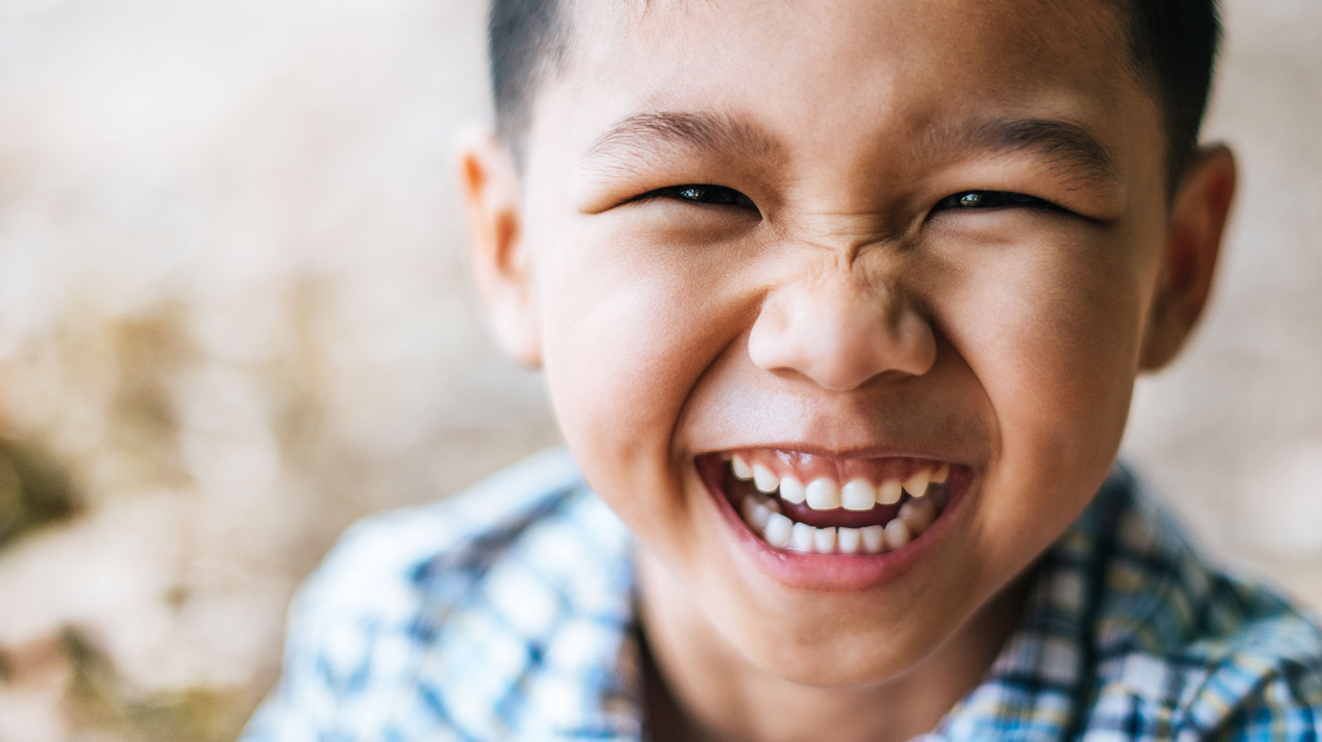 The image shows a young boy with a joyful expression, possibly laughing, smiling at the camera with his mouth open. He appears to be outdoors during daylight, wearing a light-colored shirt and dark pants, and has short hair.