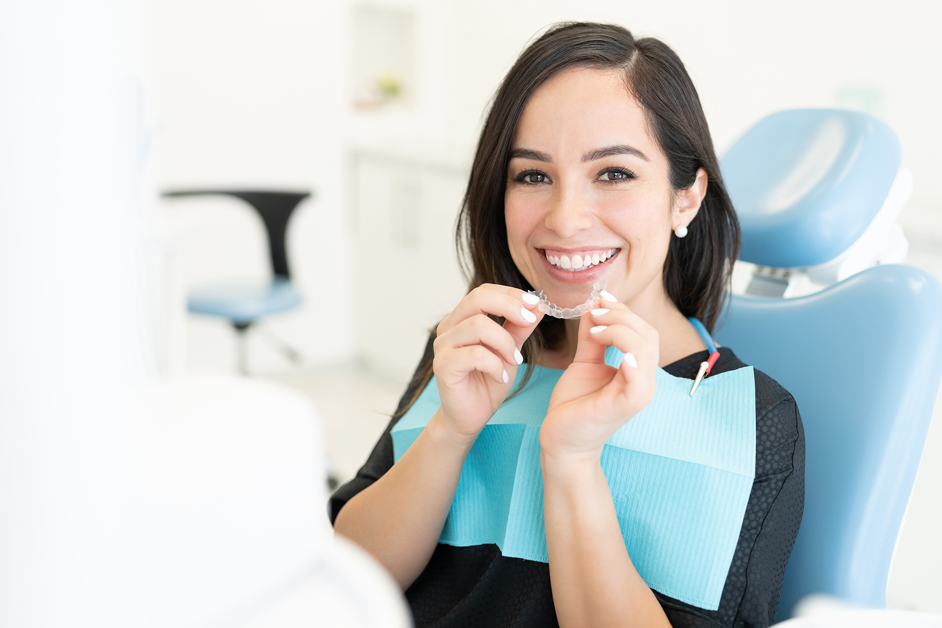 A woman sitting at a dental chair, holding a toothbrush with a smile, wearing an apron, in a dental office setting.