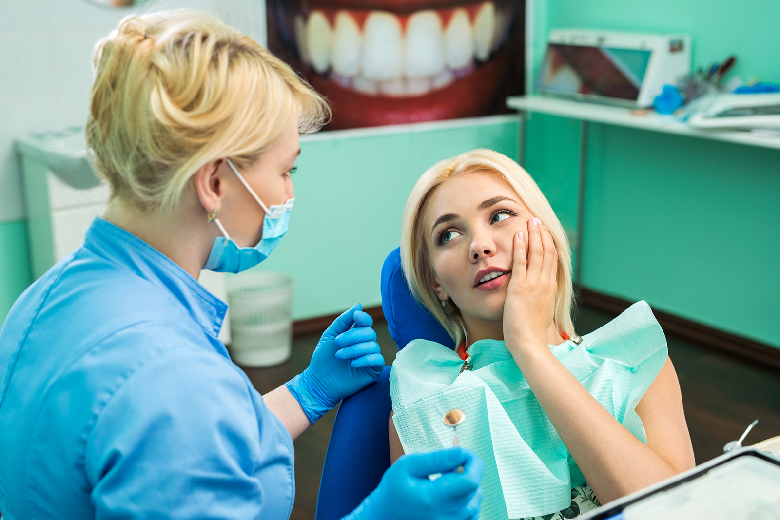 The image shows a dental office setting with a woman seated in a dental chair receiving dental care from a dentist, who appears to be cleaning her teeth.