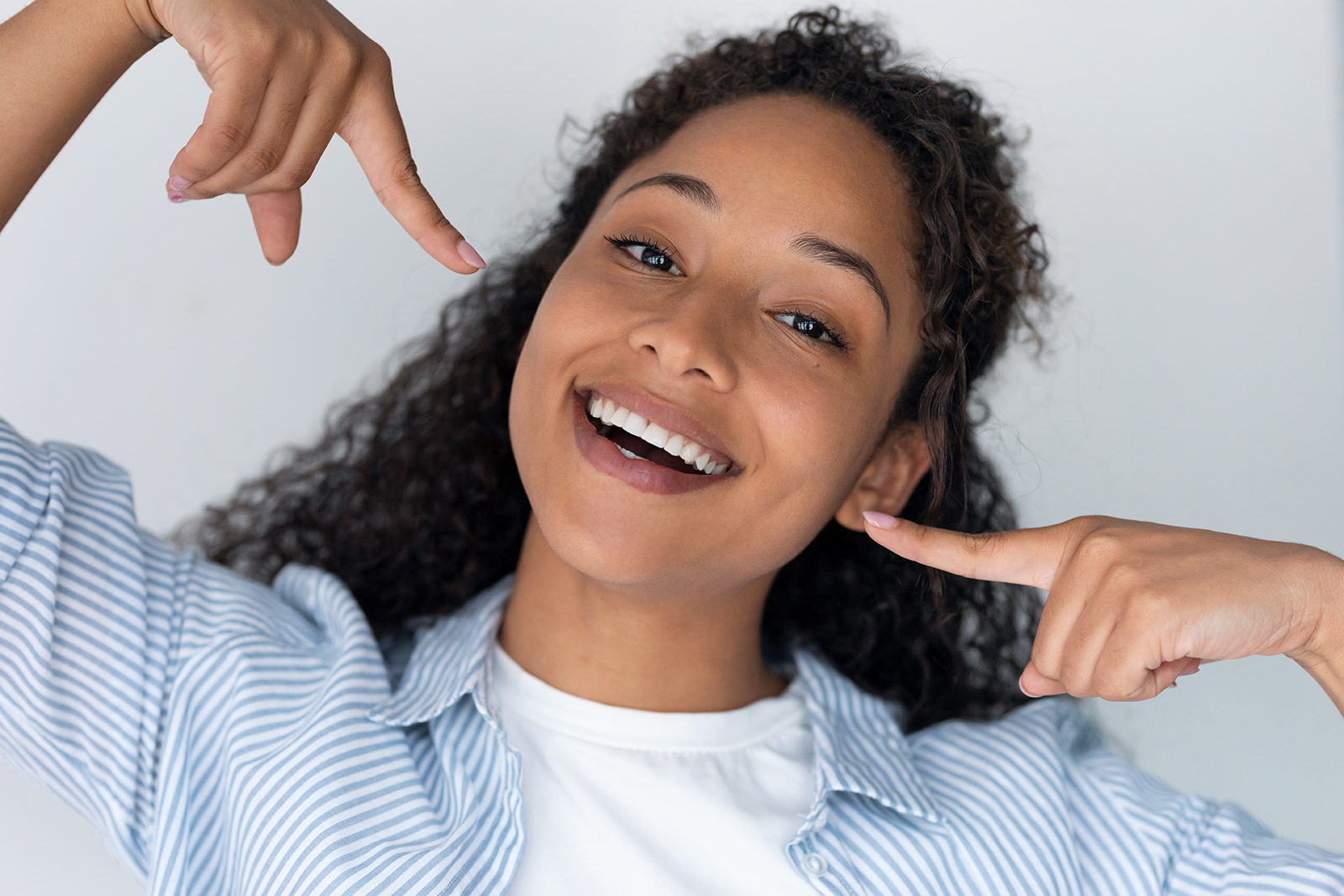 Woman with curly hair smiling broadly, making a peace sign gesture with her hands, against a white background.