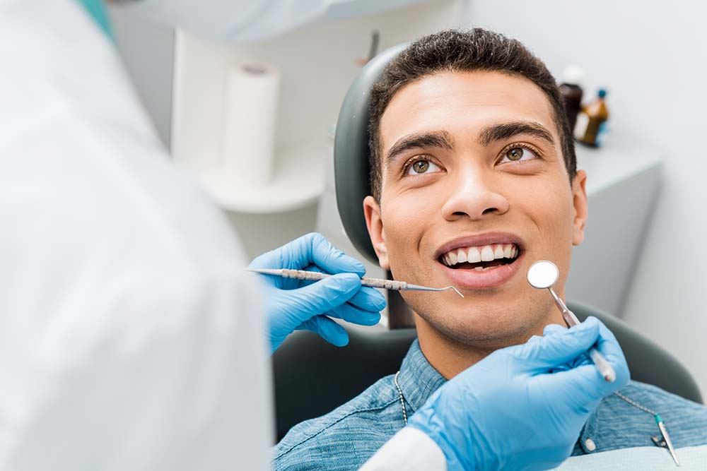 A man sitting in a dental chair with a bright smile, receiving dental care from a professional wearing protective gloves and a face mask.