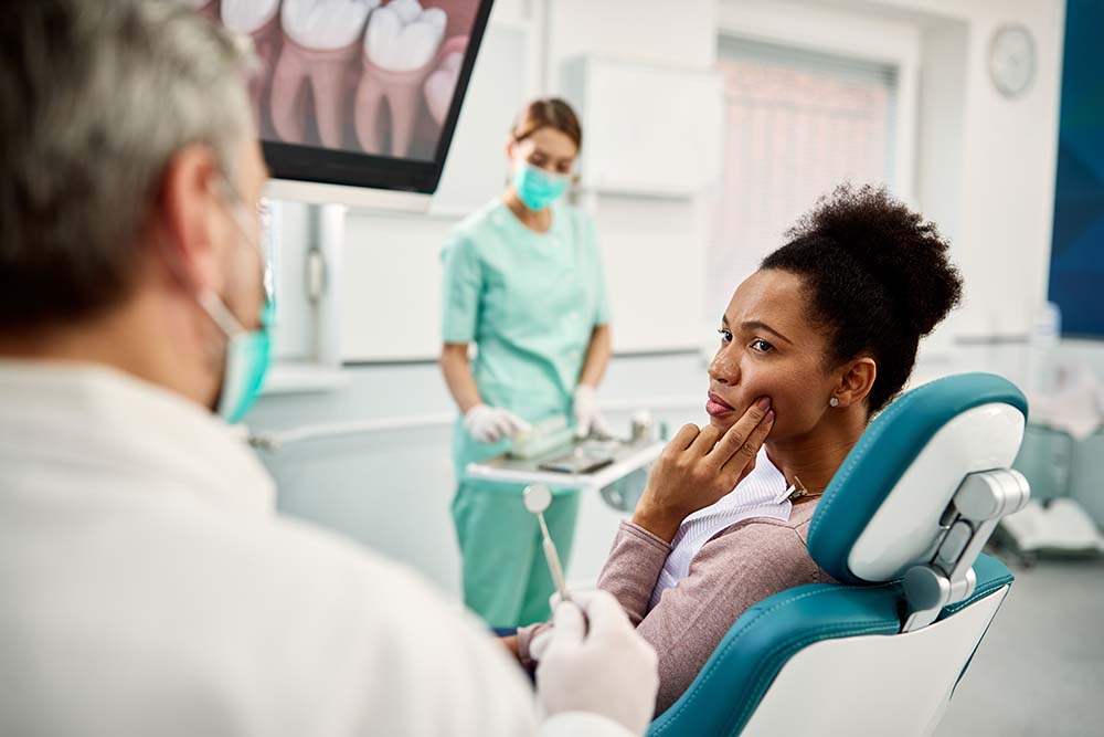 A woman sits in a dental chair with her hand on her face, receiving dental care from a dentist who appears to be examining her teeth.