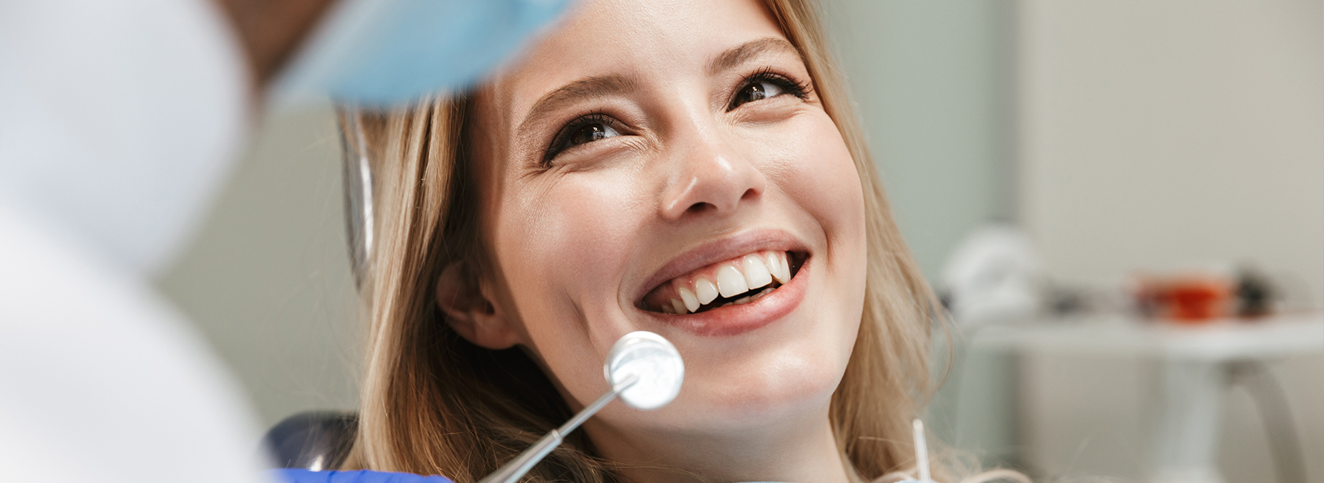 A young woman with a big smile sits in a dental chair, receiving dental care from a professional.