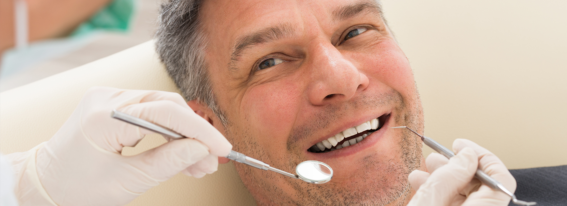 The image shows a man seated in a dental chair, receiving dental care with his mouth open, while holding a toothbrush in his hand, and wearing a white lab coat.