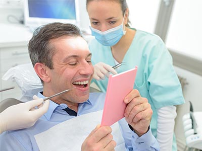 The image shows a man seated at a dental chair, looking at a pink card with a surprised expression while surrounded by dental professionals who are smiling and observing his reaction.