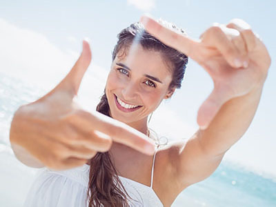 A young woman with long hair is smiling at the camera while holding up her hand to frame a picture of herself against a sunny beach backdrop.